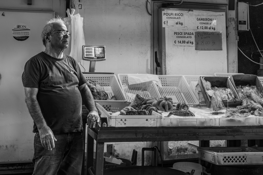 Fishmonger standing beside a seafood counter with fish and price signs.