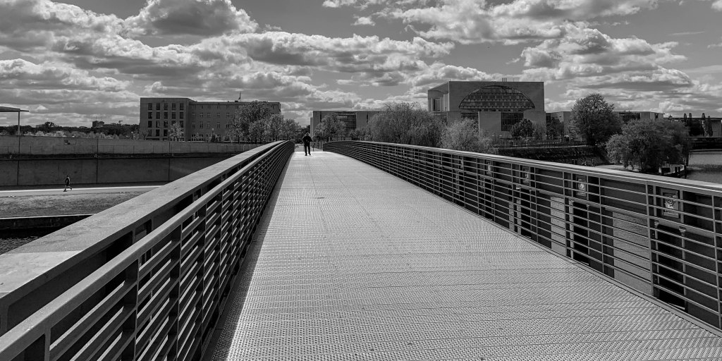 Empty pedestrian bridge in Berlin during lockdown, with railings, clouds, and distant buildings.