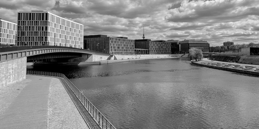 Wide view of the Spree in Berlin with modern buildings, bridge, railings, and heavy clouds.