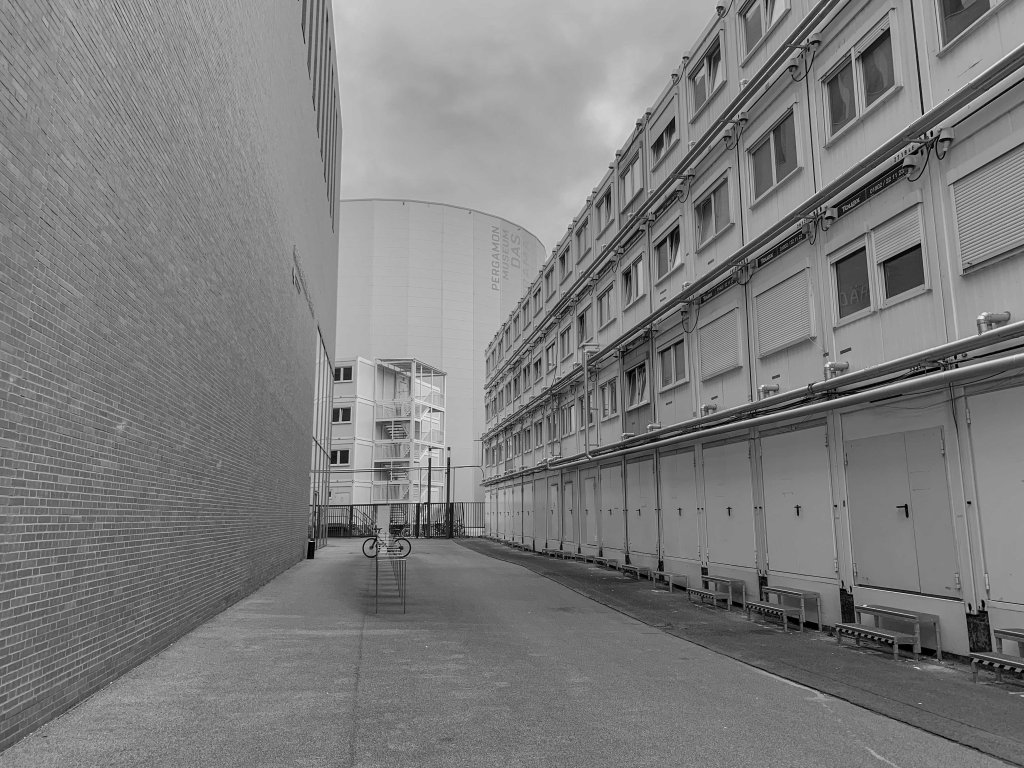 Empty urban passage between a high wall and a long building, with a bicycle rack in the distance.