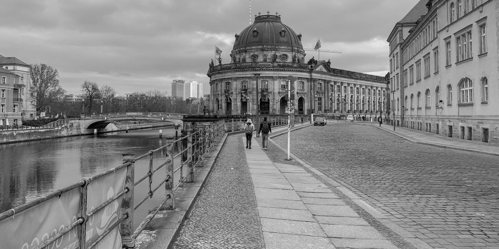 Riverside walkway leading towards the Bode Museum, with water, railings, cobblestones, and few pedestrians.