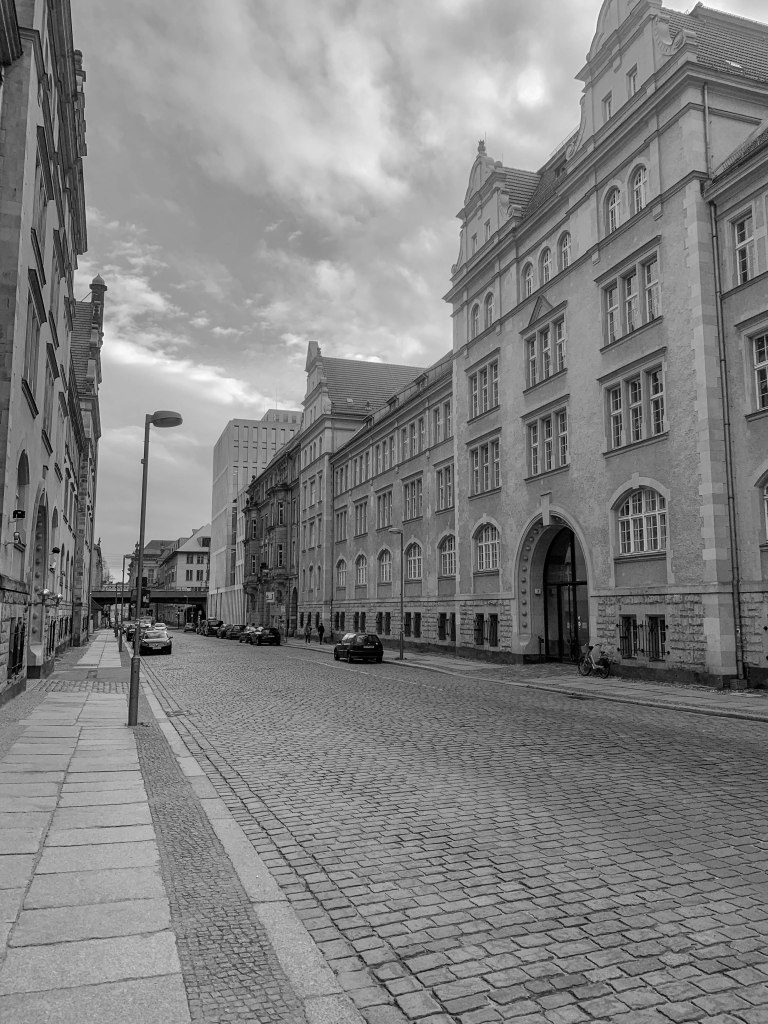 Empty cobbled Berlin street lined with large historic buildings beneath a clouded sky.