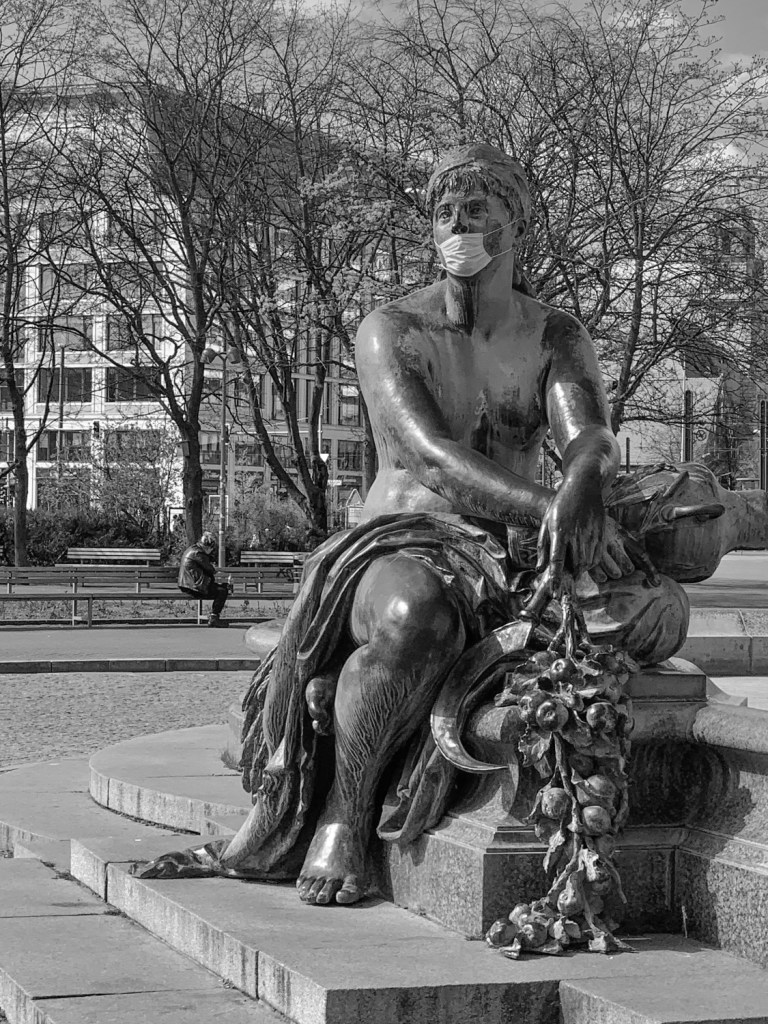 Berlin statue wearing a face mask during lockdown, seated among bare trees.