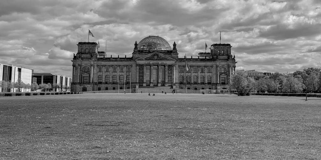 Reichstag building seen across a wide empty lawn under dramatic clouds.