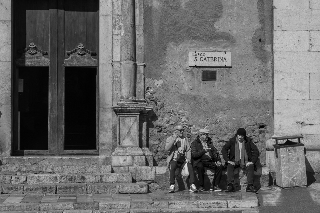 Three people sitting together against a worn wall beneath a sign reading “Largo Santa Caterina” in Taormina.