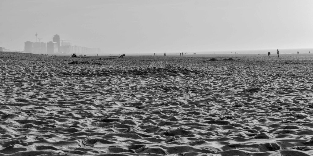 Low view across rippled sand on the Belgian coast, with distant figures and hazy buildings on the horizon.