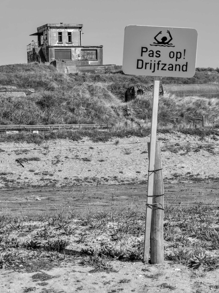 Belgian coast warning sign reading “Pas op! Drijfzand”, with dunes and an abandoned-looking building behind it.