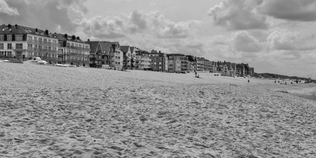 Wide beach on the Belgian coast with seafront buildings, sand, and clouds above the promenade.