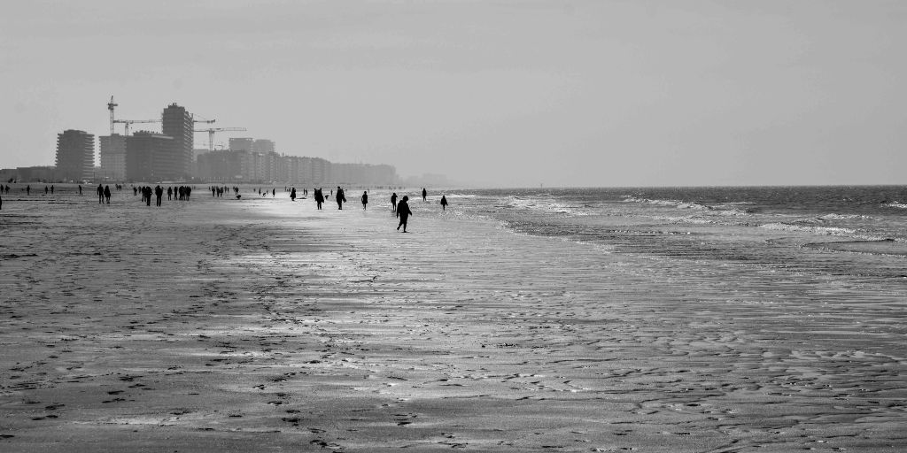 Long shoreline view with small figures walking on wet sand beside the sea and distant buildings.