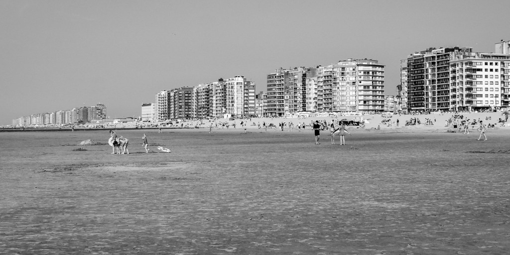 Broad Belgian beach with scattered people and a long line of modern seafront apartment blocks.