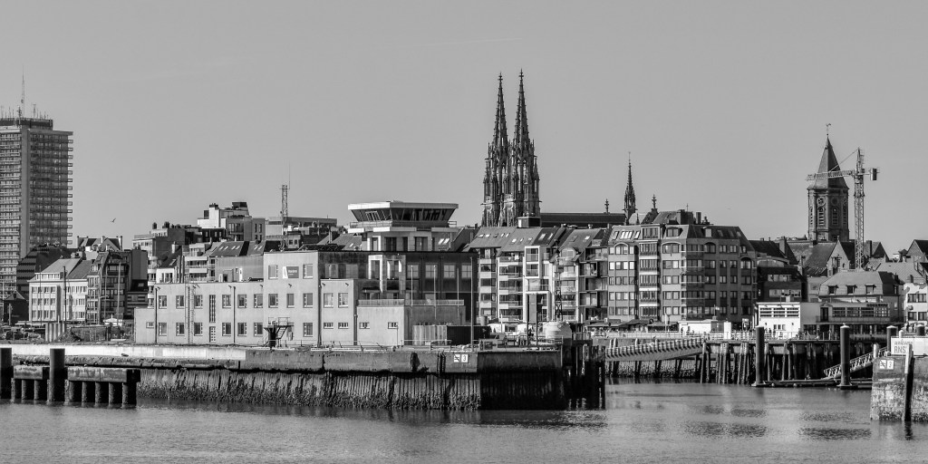 Harbour view of Ostende with buildings, church towers, piers, and water in the foreground.