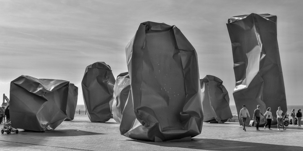 Large folded metal sculptures on a Belgian seafront square, with people walking nearby.