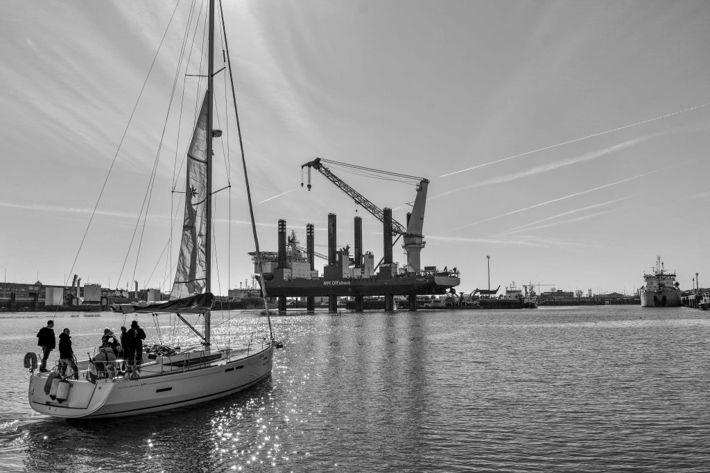 Sailboat on the water near Ostende harbour, with cranes, industrial structures, and sunlight on the sea.