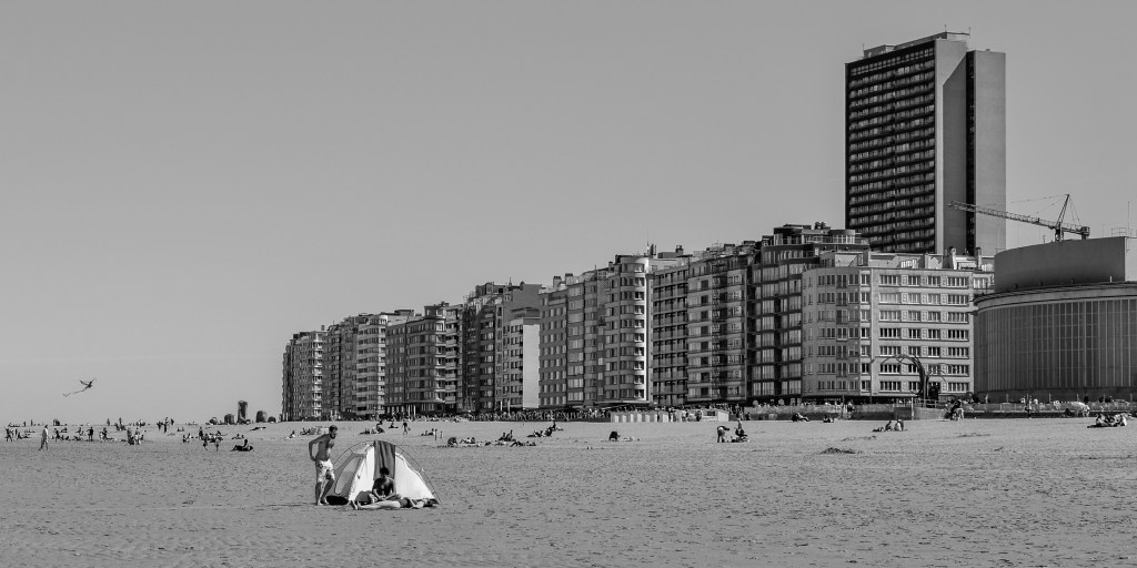 Belgian beach with tall seafront buildings, scattered figures, tents, and open sand.