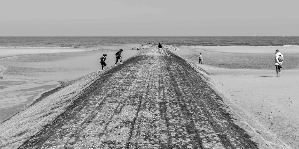 Long groyne or breakwater extending towards the sea, with people walking along it and wet sand on both sides.
