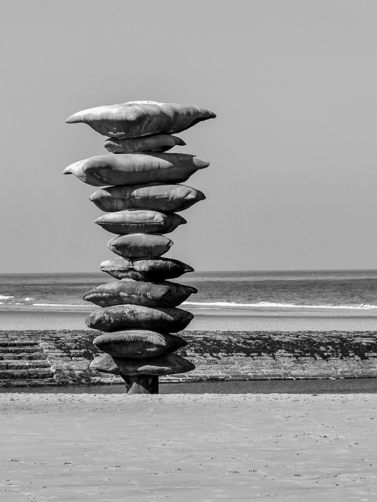 Stacked sculptural form on the beach, with the sea and groynes behind it.