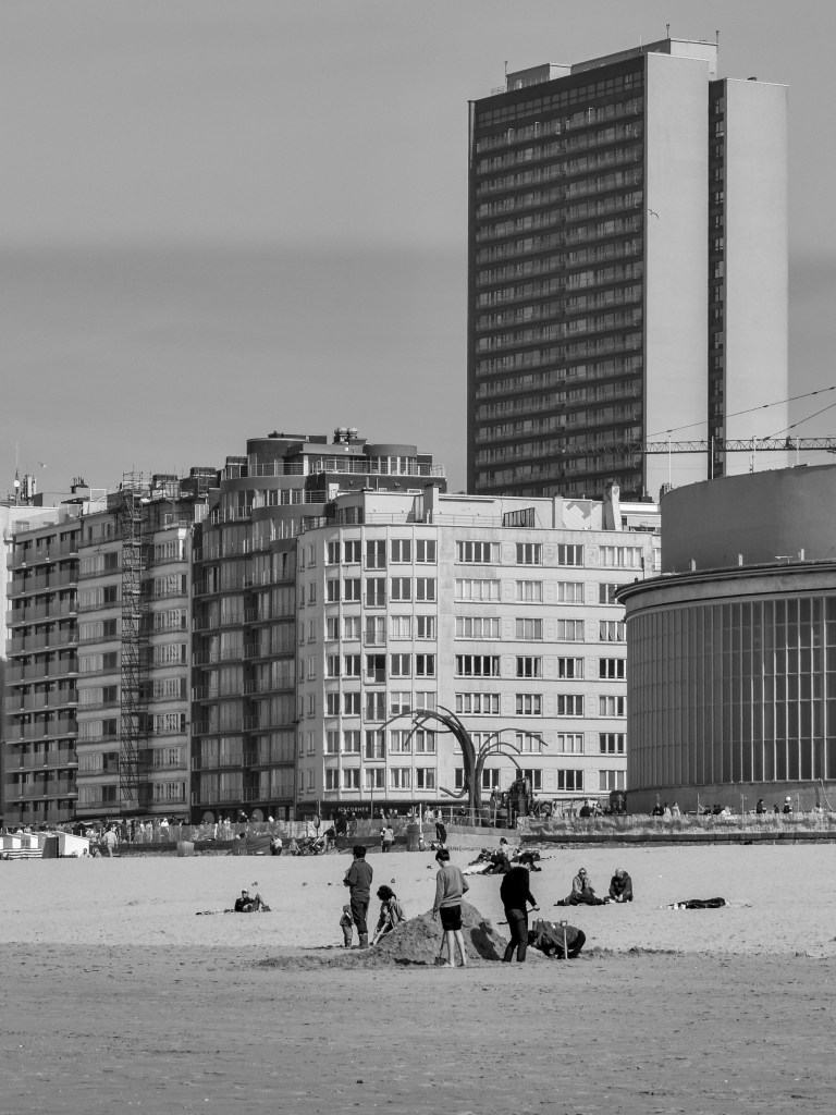 Ostende beach with walkers and sunbathers in front of tall apartment buildings and a large tower.