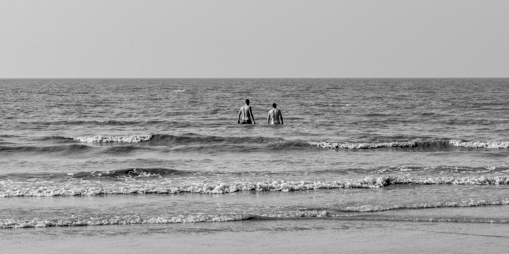 Two figures standing in shallow sea water on the Belgian coast, with small waves crossing the frame.