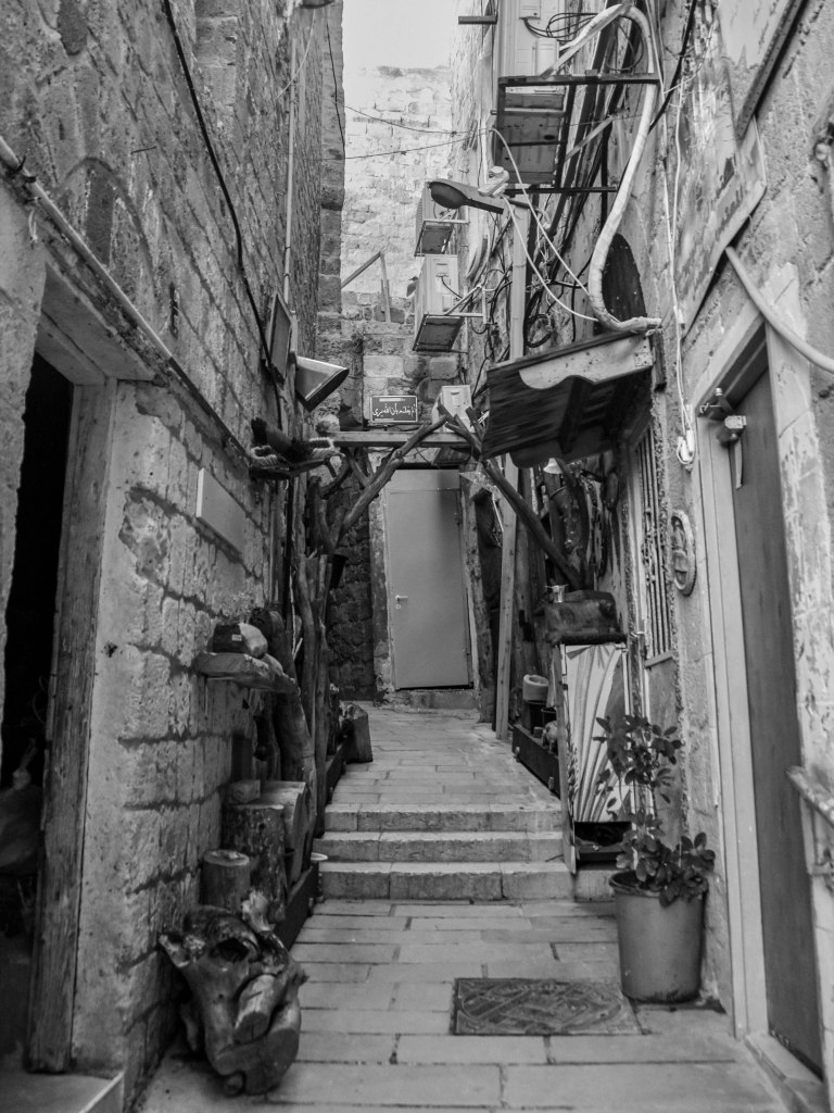 Stone alley with steps, pipes, plants, doors, and rough walls in an old city.