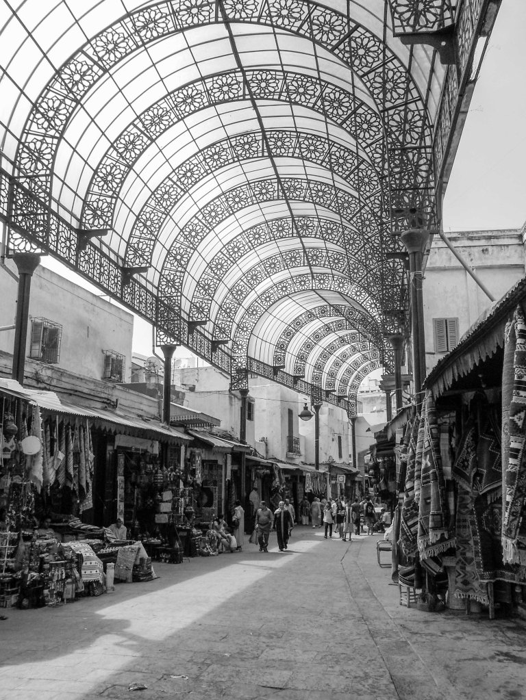 Covered market passage with arched metal roof, stalls, pedestrians, and light filtering through the structure.