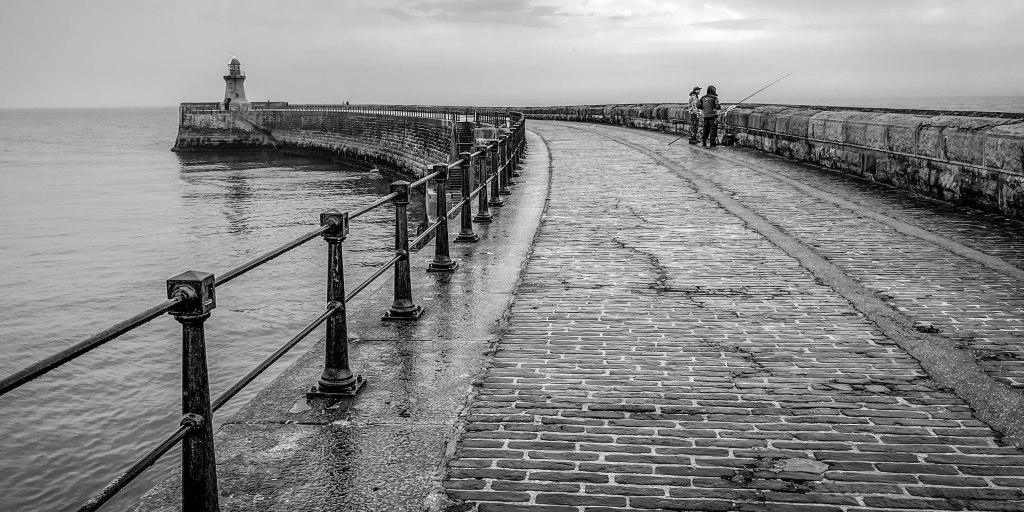 Wet stone pier curving into the sea, with railings, lighthouse, and figures near the end.