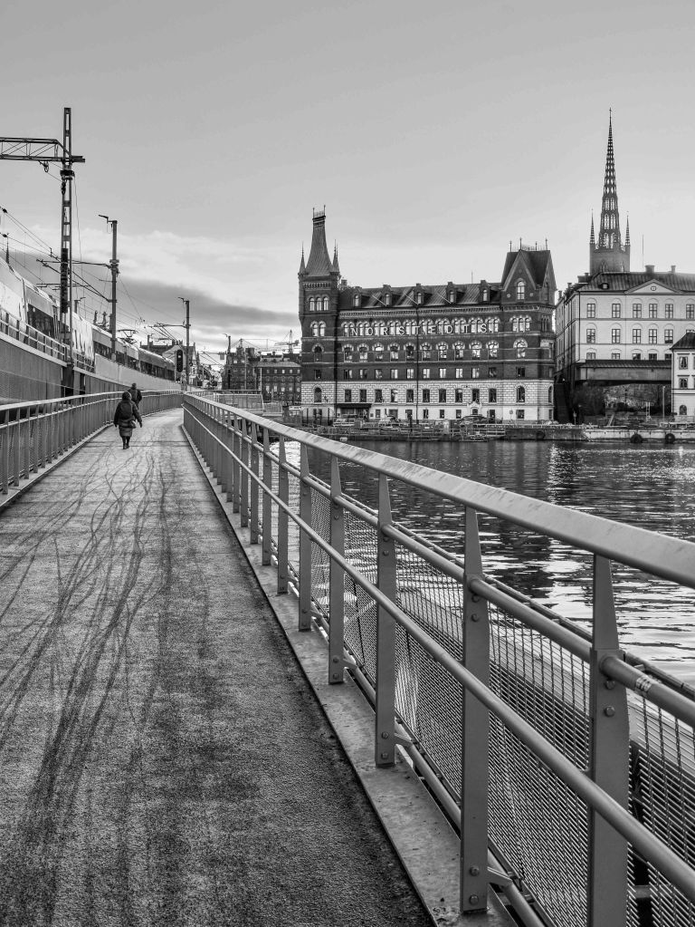 Riverside walkway with railings, water, historic buildings, and a distant church spire.