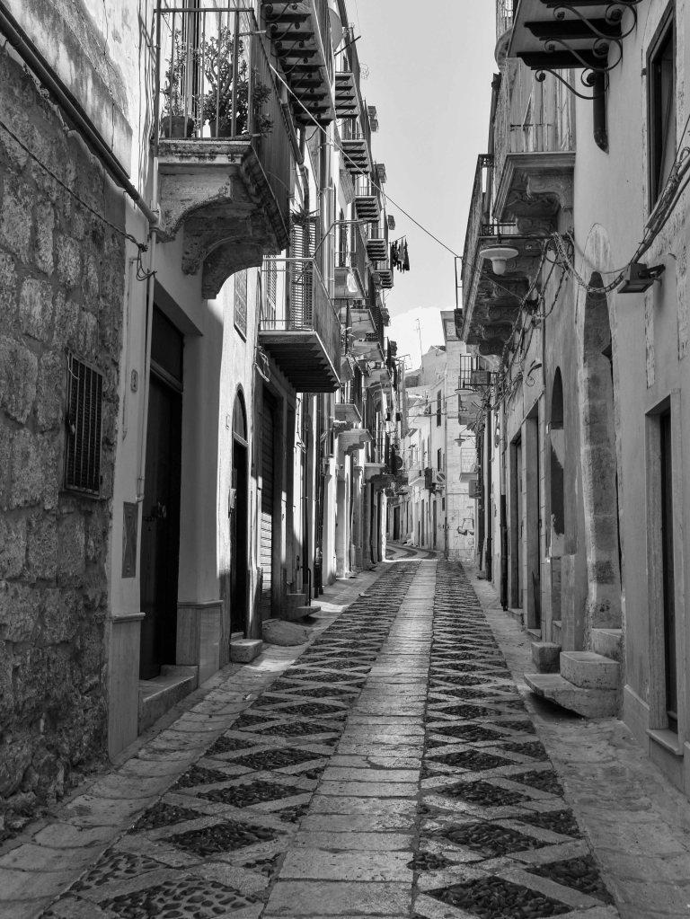Narrow stone street with patterned paving, balconies, doors, and bright sunlight.