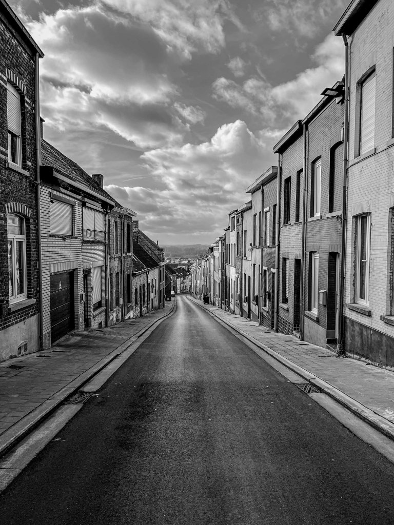 Steep residential street descending between houses towards a distant view beneath a clouded sky.