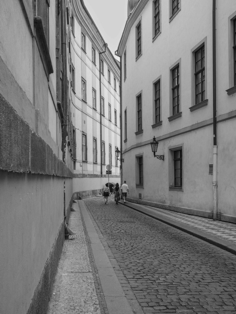 Quiet cobbled street in Prague between pale buildings, with two distant figures walking away.