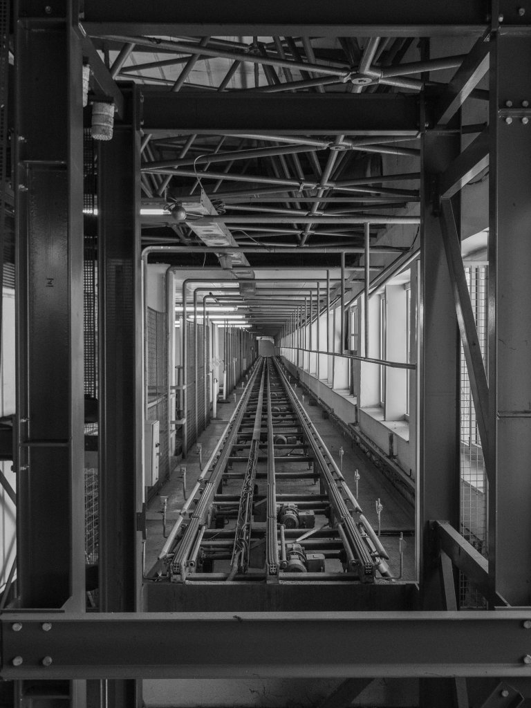 Industrial passage with rails, pipes, beams, and metal frames receding into the distance.