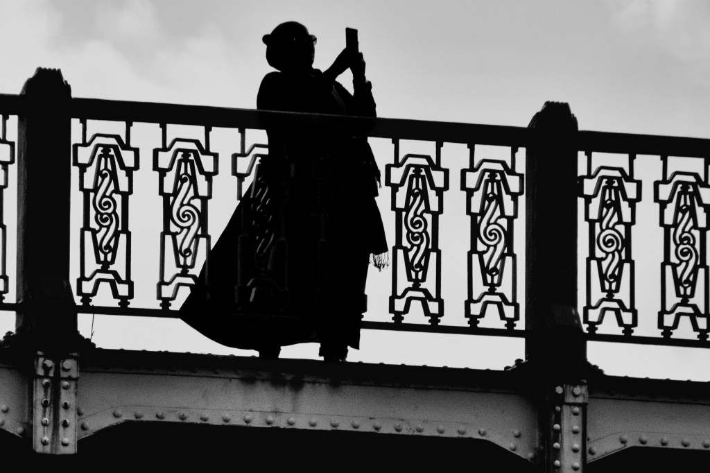 Silhouetted figure crossing a bridge with a bag, framed against an ornamental railing.
