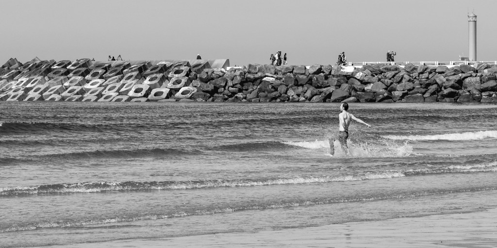 Person running across shallow water on a Belgian beach near a rocky breakwater.