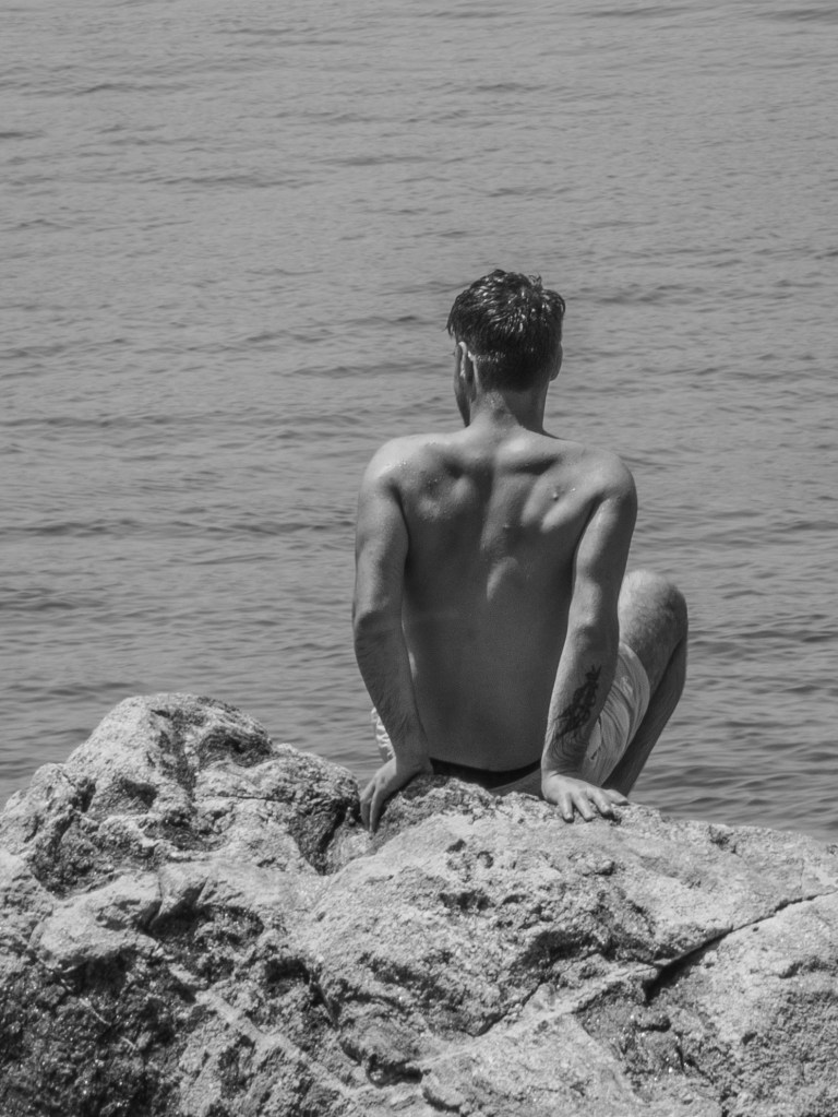 Shirtless man sitting on rocks and looking out over the sea.