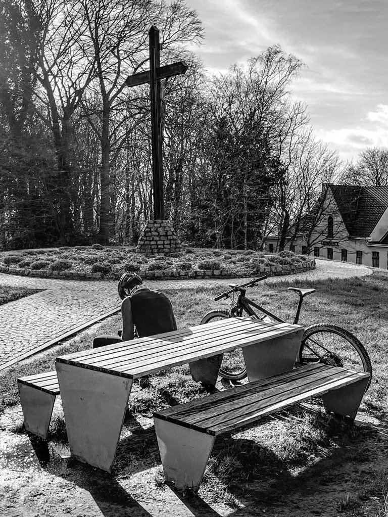 Person sitting near a bench and bicycle in a park, with a large cross behind them.