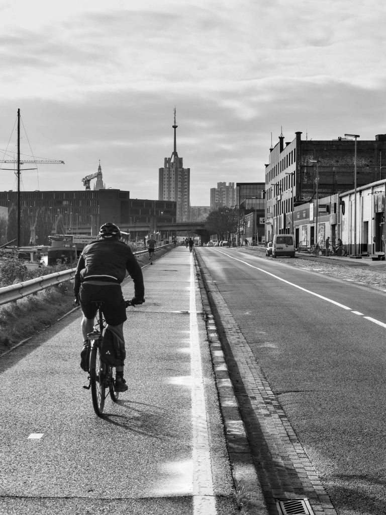 Cyclist riding along a broad urban road beside industrial buildings.