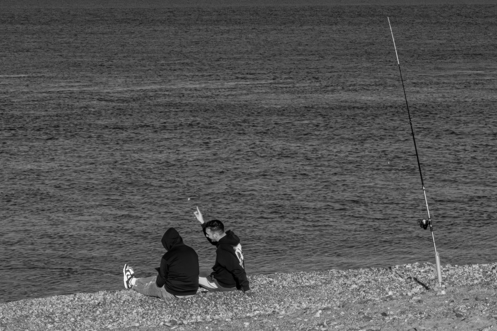 Two people sitting on a beach beside a fishing rod, looking towards the sea.