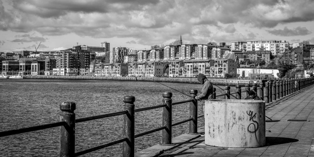 Person resting beside a waterfront railing, with a city across the water.