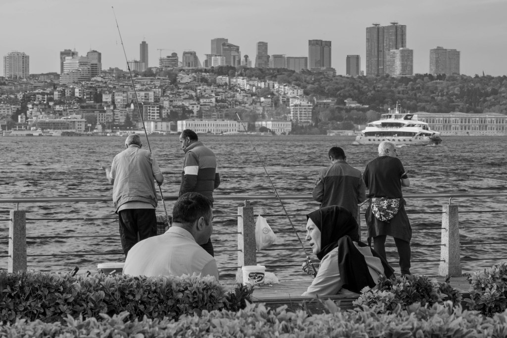 People resting and fishing by the water, with Istanbul’s urban shoreline behind them.