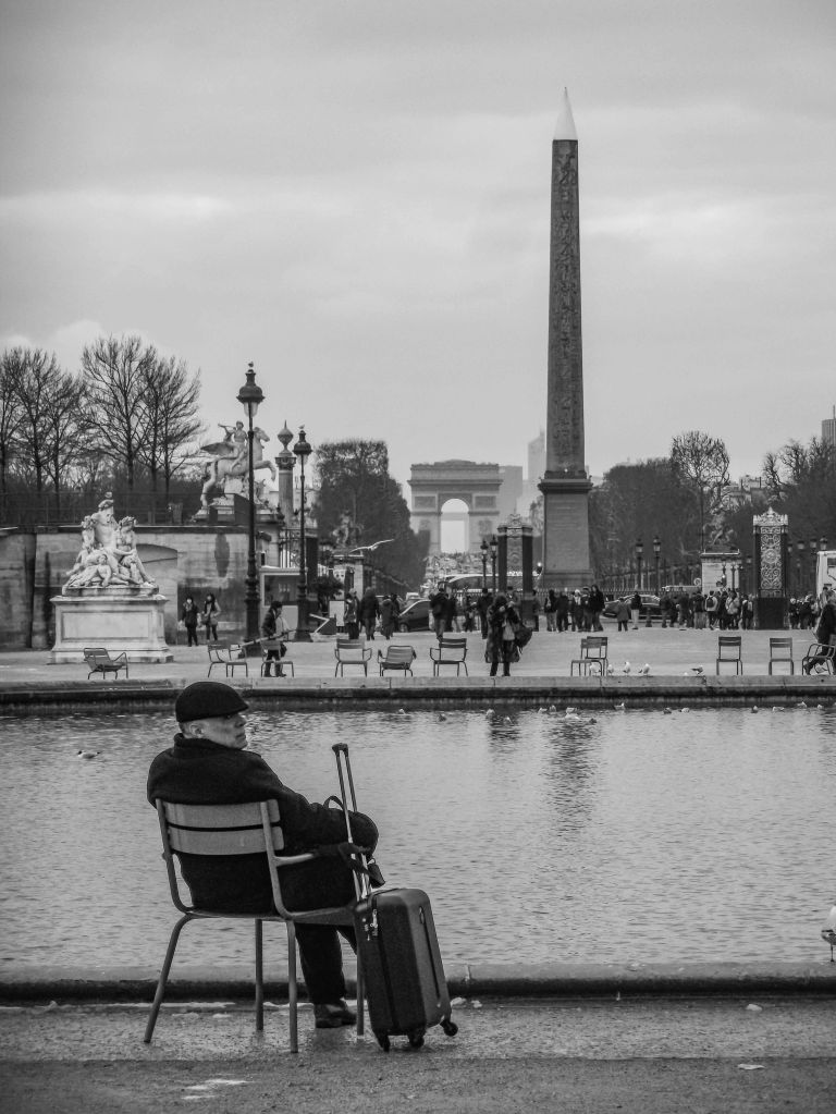 Person sitting alone on a chair beside water in Paris, with the Luxor Obelisk and Arc de Triomphe in the distance.