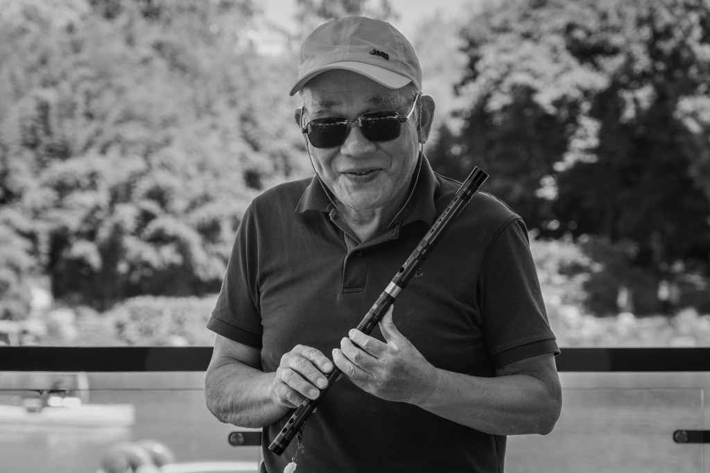 Older man smiling while playing a flute outdoors.