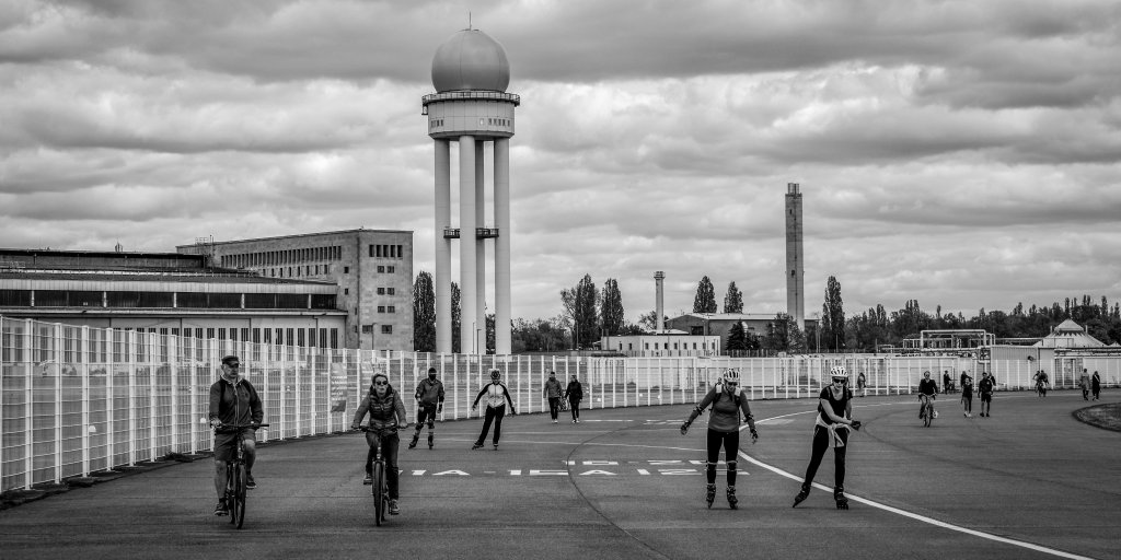 People skating or cycling in a wide public square under a cloudy sky.