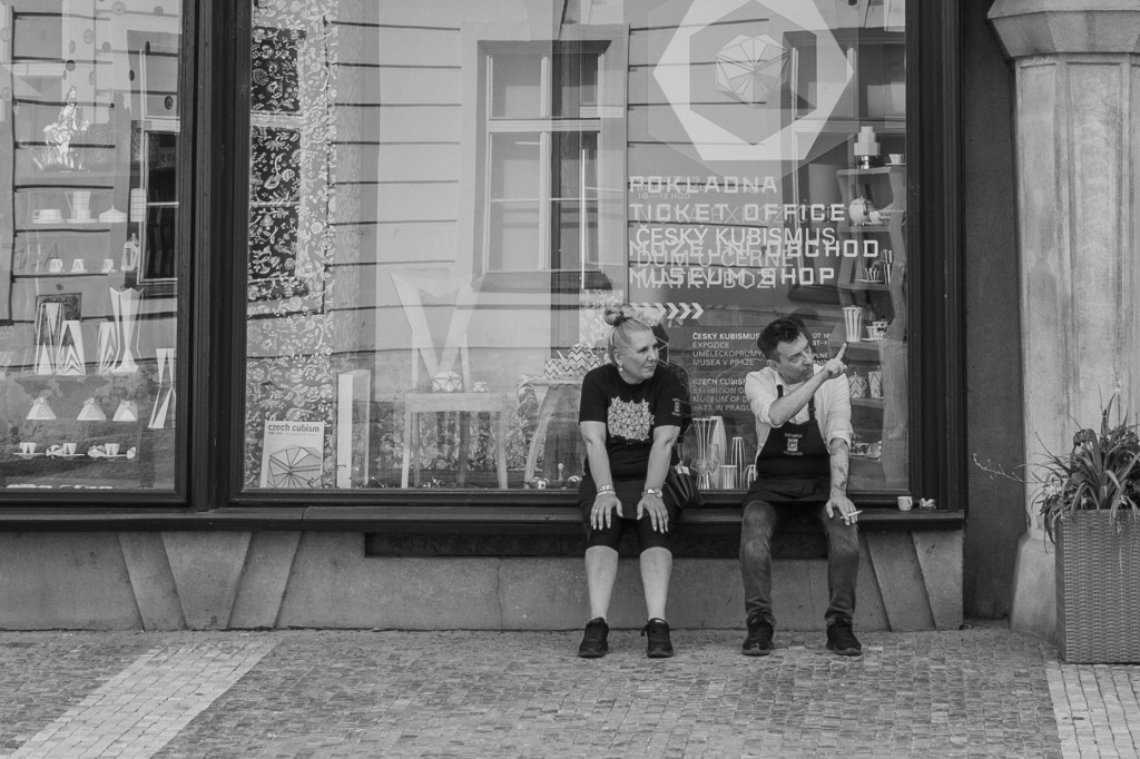 Two people sitting outside a shop window, resting in front of reflected street text.