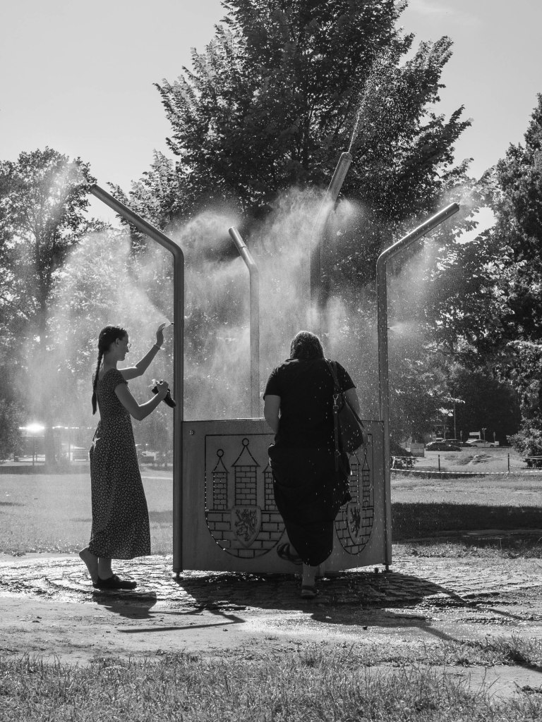 Two people using a public misting fountain in a park, with spray lit from behind.