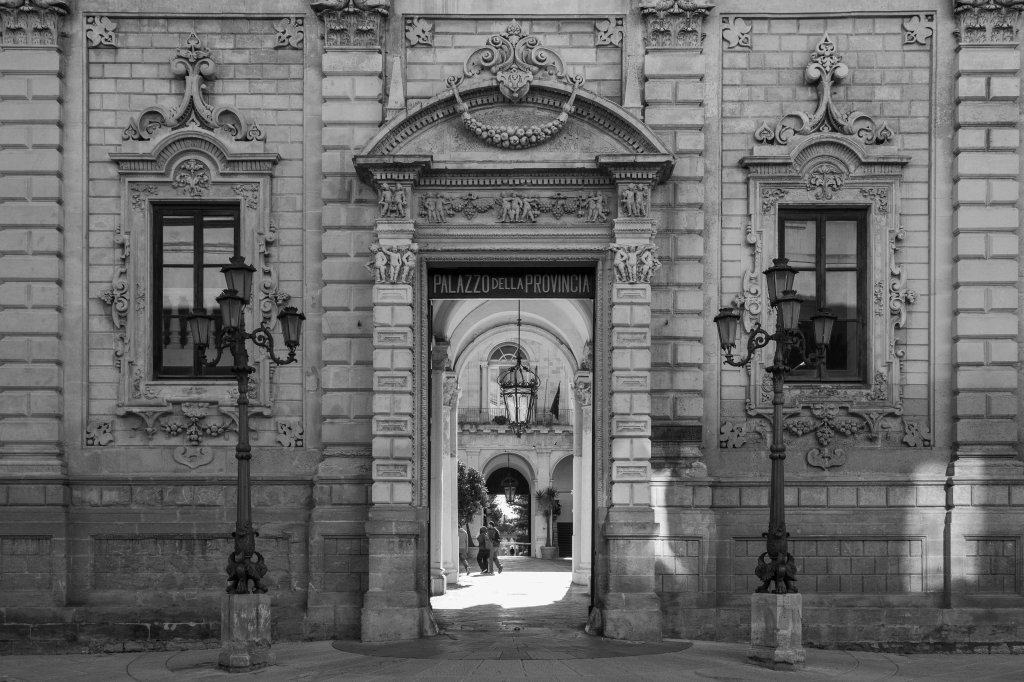 Ornate stone façade with central arched portal, decorated windows, lamps, and a passage visible through the entrance.