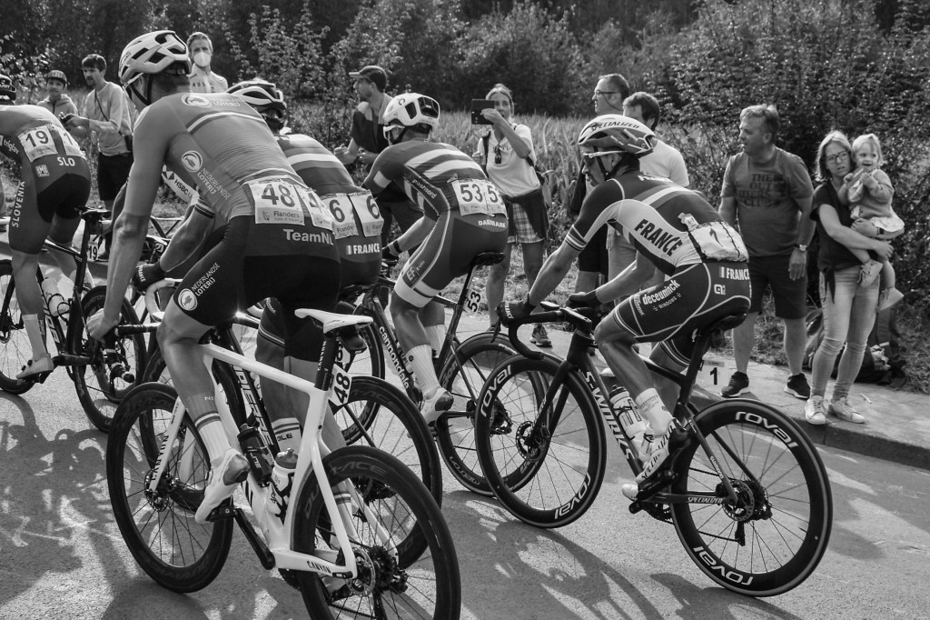 Cyclists riding close together, seen from behind and beside, with spectators watching from the roadside.