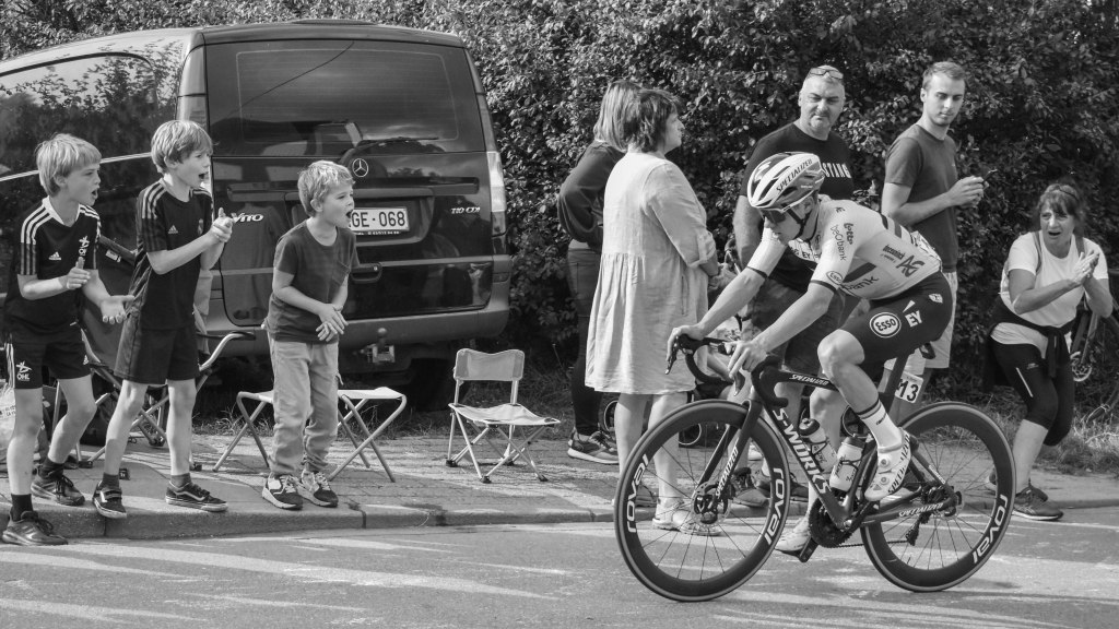 Lone cyclist passing children and spectators cheering from the side of the road.