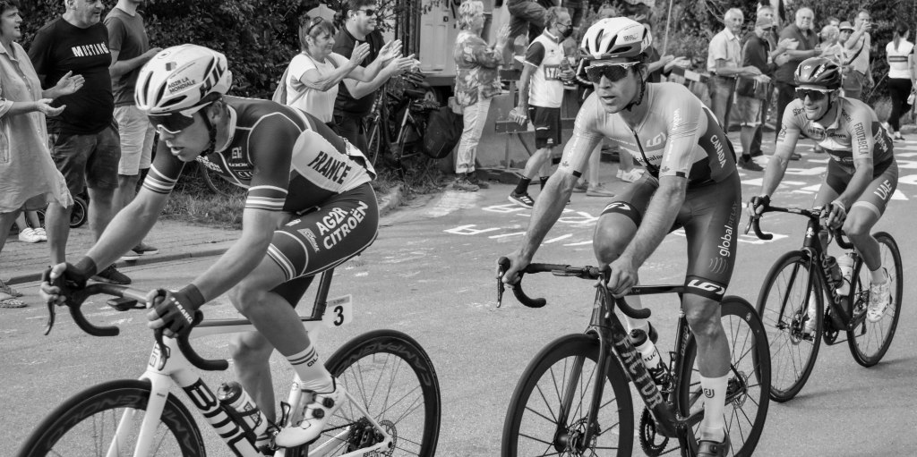 Small group of cyclists riding in line, with spectators and barriers behind them.