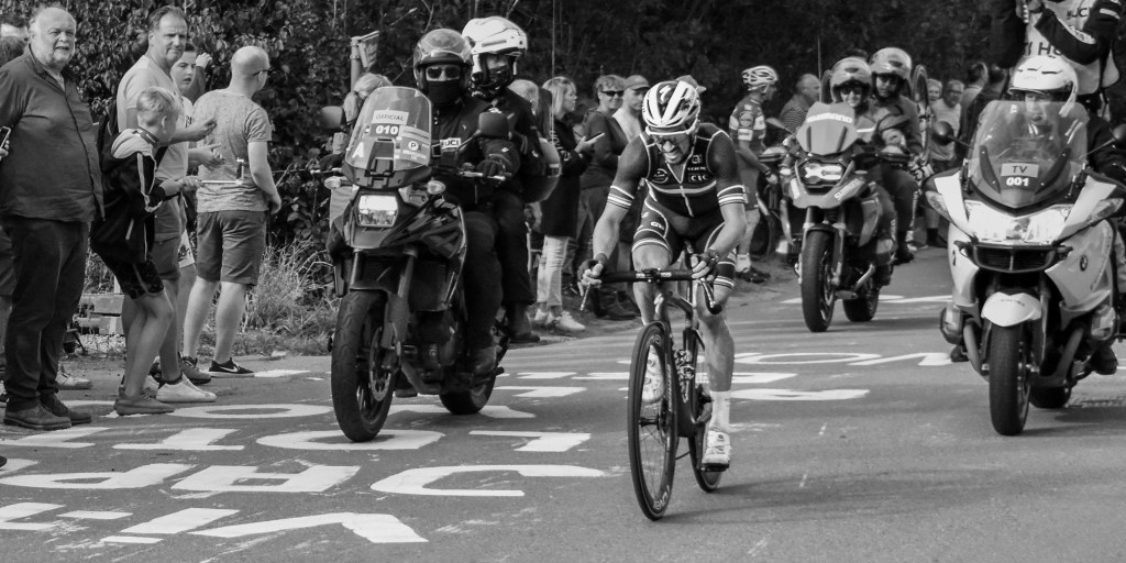 Cyclist riding ahead of motorbikes and race officials, with spectators along the road.