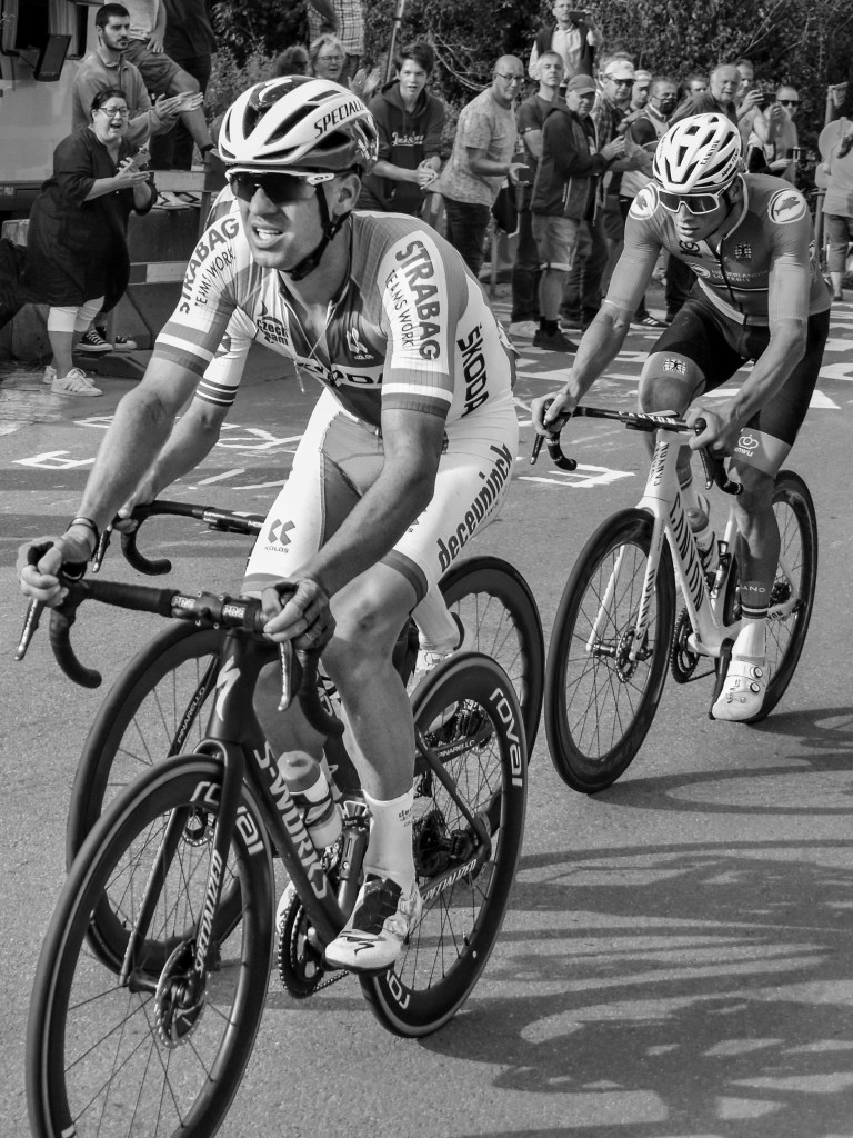 Two cyclists riding closely together, one in a white kit in the foreground and another following behind.