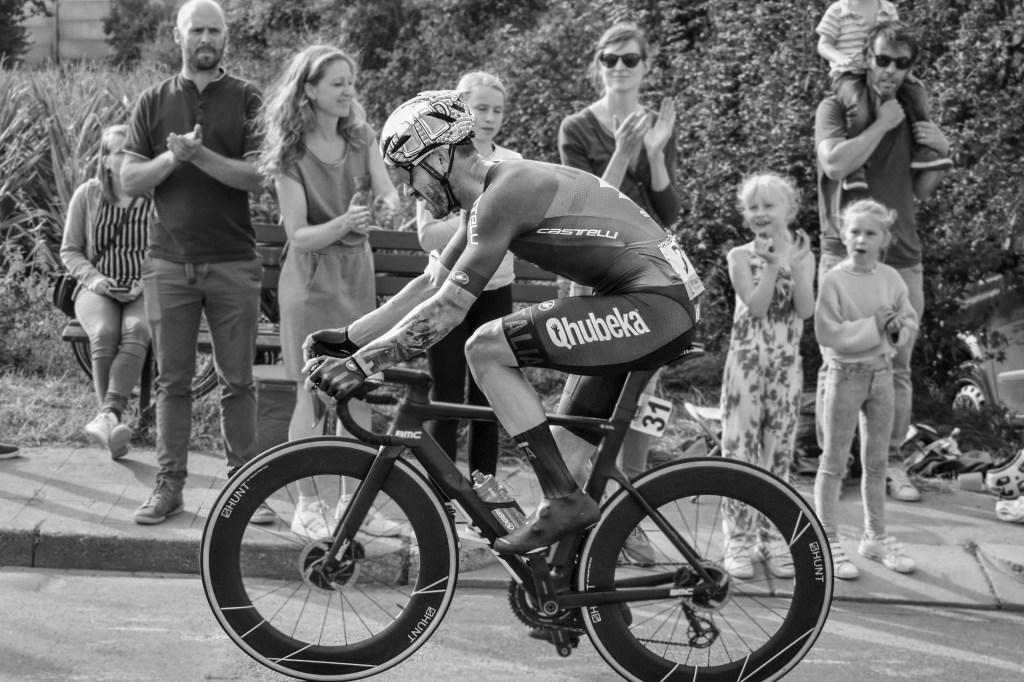 Lone cyclist passing a family of spectators, with children and adults watching from the roadside.